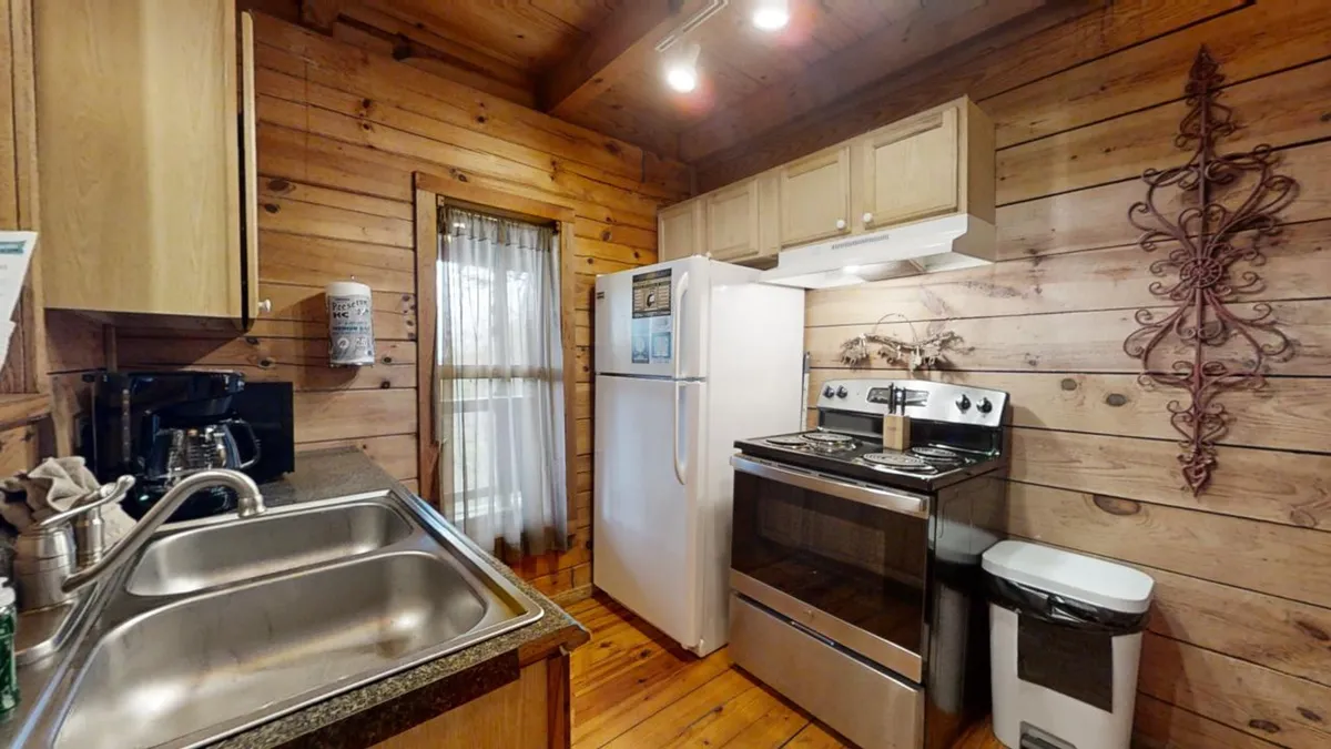 A warm, homely kitchen featuring a kitchen sink, stove, refrigerator, wooden walls, and curtains. The setting evokes a sense of togetherness and is designed for cooking, gathering, and meal preparation, with modern appliances and ample counter space evident.