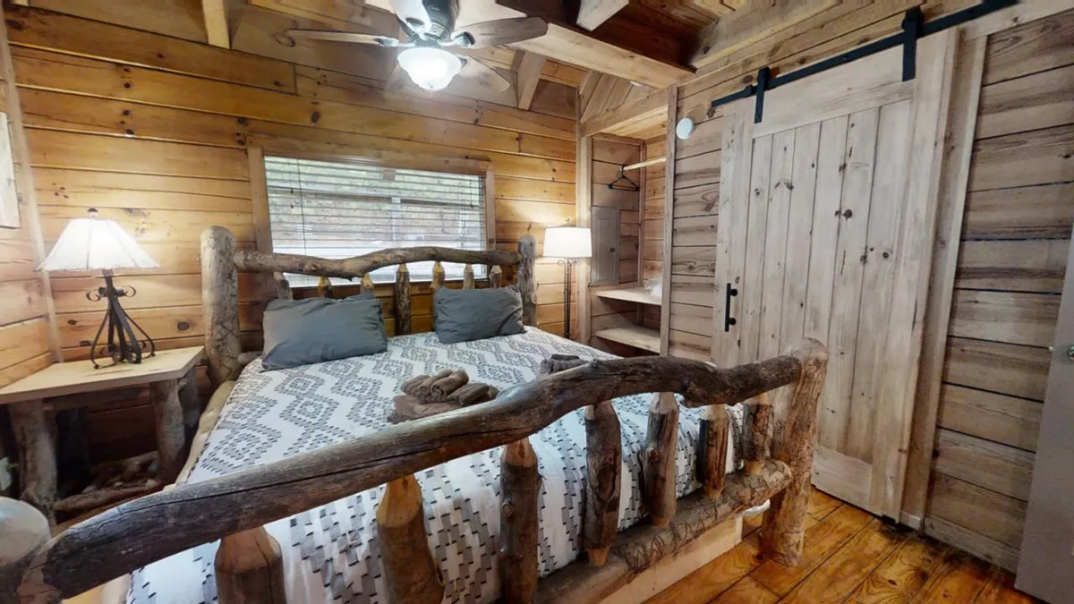 A warmly lit bedroom featuring a rustic wooden bed frame, a decorative throw draped over the foot of the bed, a table lamp casting a soft glow, and large window letting in natural light. The space exudes rustic charm while emphasizing comfort and a connection to nature.