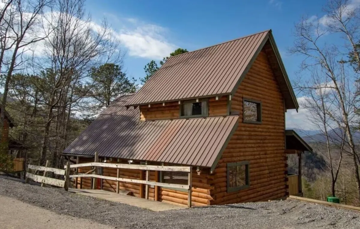 This image shows a cozy log cabin with a sloped metal roof and a spacious deck area surrounded by natural greenery. The deck is ideal for outdoor relaxation and socializing, with ample space for seating and enjoying the views. The overall scene evokes a sense of tranquility and connection to the outdoors, perfectly capturing the spirit of mountain living.