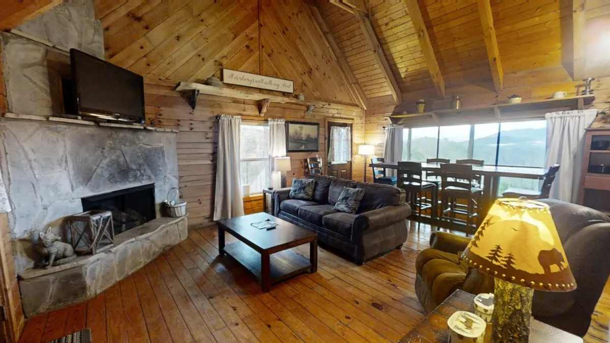 A warm, welcoming living room featuring a beautiful stone fireplace as the centerpiece, flanked by a plush sofa and large windows offering sweeping views of the mountains. The dining area with high stools is seamlessly connected, and the cozy ambiance is enhanced by decorative lighting.