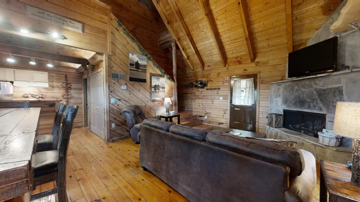 A wide shot of a warm and welcoming living area featuring exposed wooden beams, a cozy stone fireplace, a comfortable sofa, and a glimpse of the dining area. The space exudes a rustic mountain charm and feels connected to the outdoors through its inviting atmosphere.