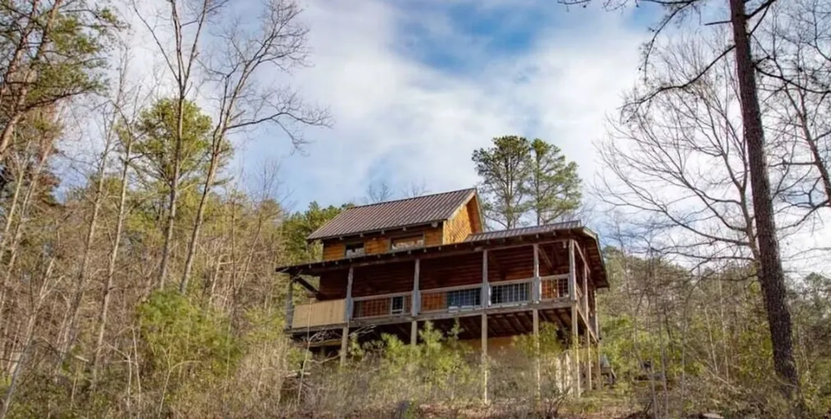 This exterior shot features the rustic wooden cabin nestled among tall trees, with a balcony structure visible. The surrounding natural landscape highlights the cabin’s secluded, adventure-ready setting, perfect for families returning from exciting nearby attractions.