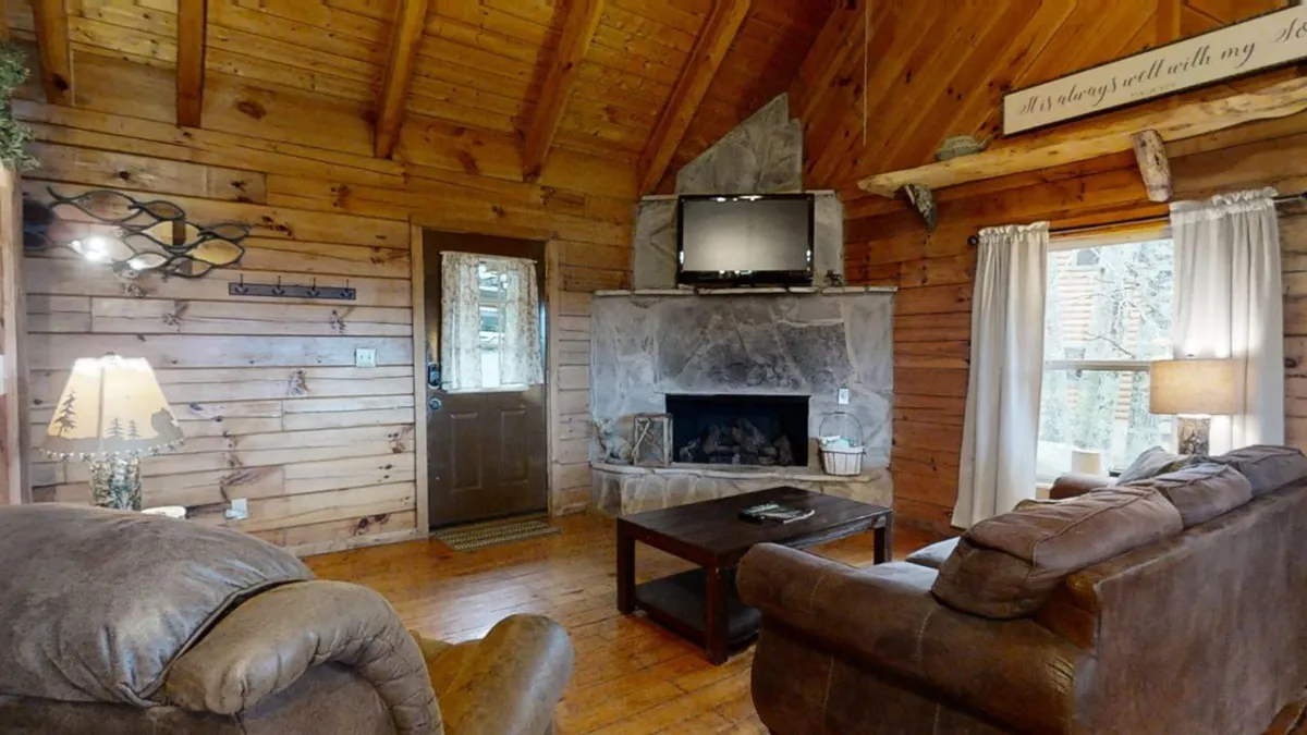 A cozy scene featuring a wooden fireplace with a TV mounted above, comfortable sofas arranged for conversation or movie nights, and an entrance door bringing in natural light. The mood is intimate, ideal for relaxing or entertaining.