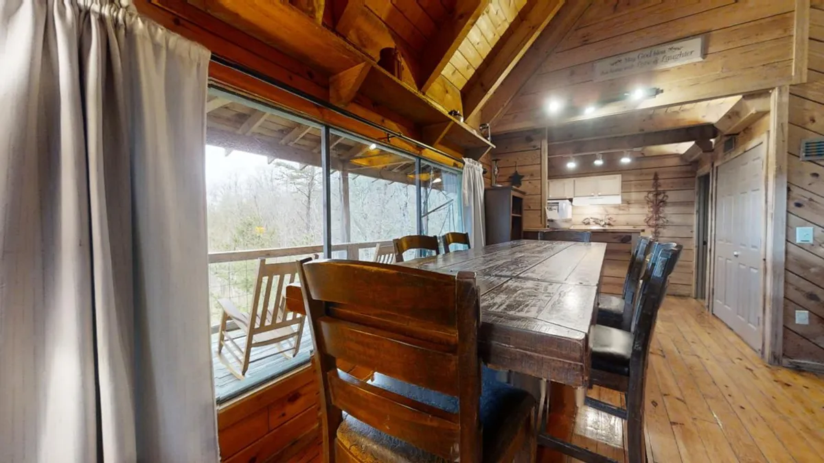 A spacious dining area with a large wooden table, surrounded by floor-to-ceiling windows that frame stunning views of nature. Wooden rocking chairs and rustic beams add to the cabin's charm, suggesting a perfect spot for shared meals or board games.