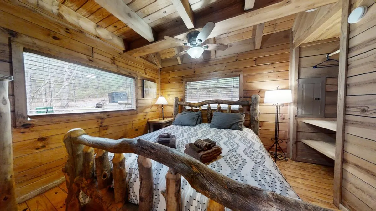 A comfortable bedroom with a log bed adorned with textured bedding, wooden walls, large windows that frame the outdoors, and a lamp for evening relaxation. The ambiance is warm and welcoming, perfect for restful nights.