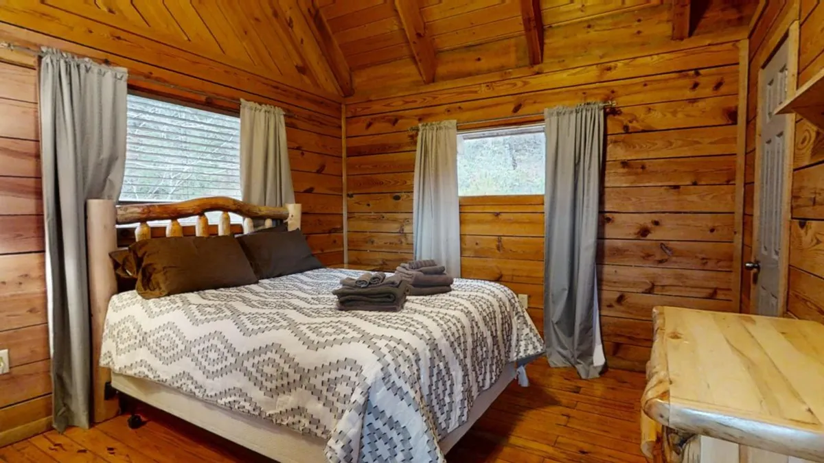 A serene bedroom with a wooden bed frame, textured bedding, large windows that open to natural light, and wood-paneled walls, creating a cozy and inviting atmosphere for rest and relaxation. The view through the windows hints at the natural beauty just outside, reinforcing the cabin's connection to the Smoky Mountain landscape.